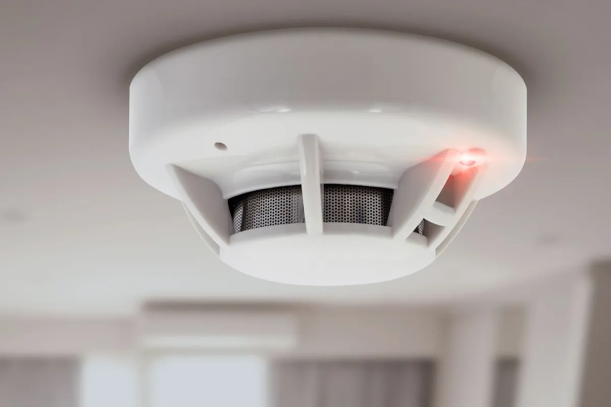 A white smoke detector mounted on a ceiling, with a red indicator light glowing