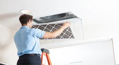 A technician in a light blue shirt replacing an HVAC air filter in a ceiling vent