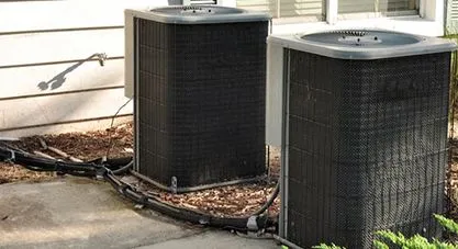 Two outdoor air conditioning units sitting on mulch next to a house
