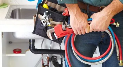 A plumber, wearing a tool belt, holding a large pipe wrench and hoses, with a kitchen sink behind him