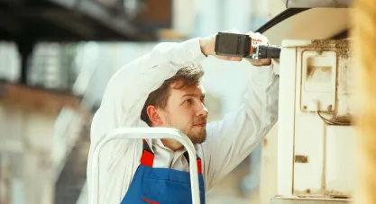 An HVAC technician on a ladder using a power drill to install an outdoor AC unit