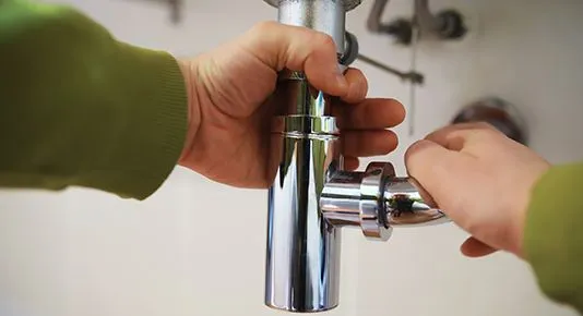 A close-up image showing hands tightening a chrome drainpipe connection under a sink