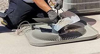 A technician performing maintenance on an outdoor AC unit, with the condenser fan assembly detached and resting on the ground