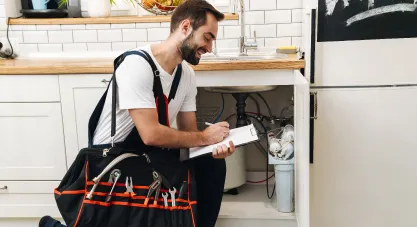 A smiling plumber inspecting a kitchen sink, writing on a clipboard