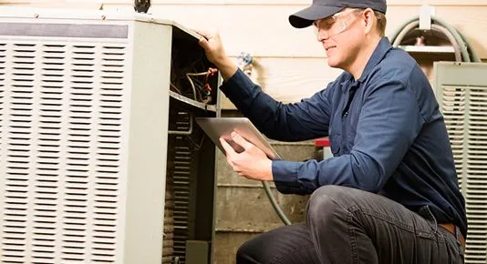 A technician in safety glasses inspecting an open AC unit, holding a tablet