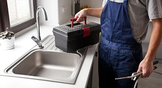 A plumber standing by a kitchen sink, holding a toolbox and plumbing tools